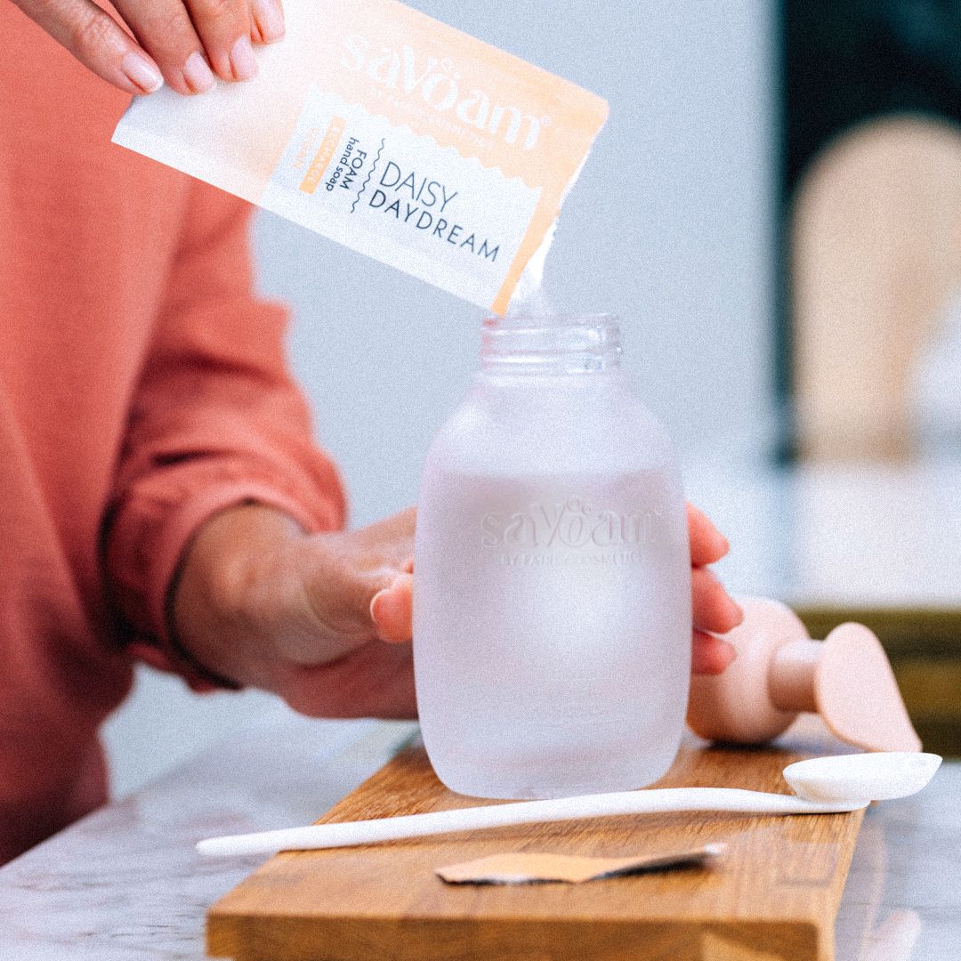 Person pouring Daisy Daydream powder into a frosted glass jar on a wooden tray with a spatula nearby.