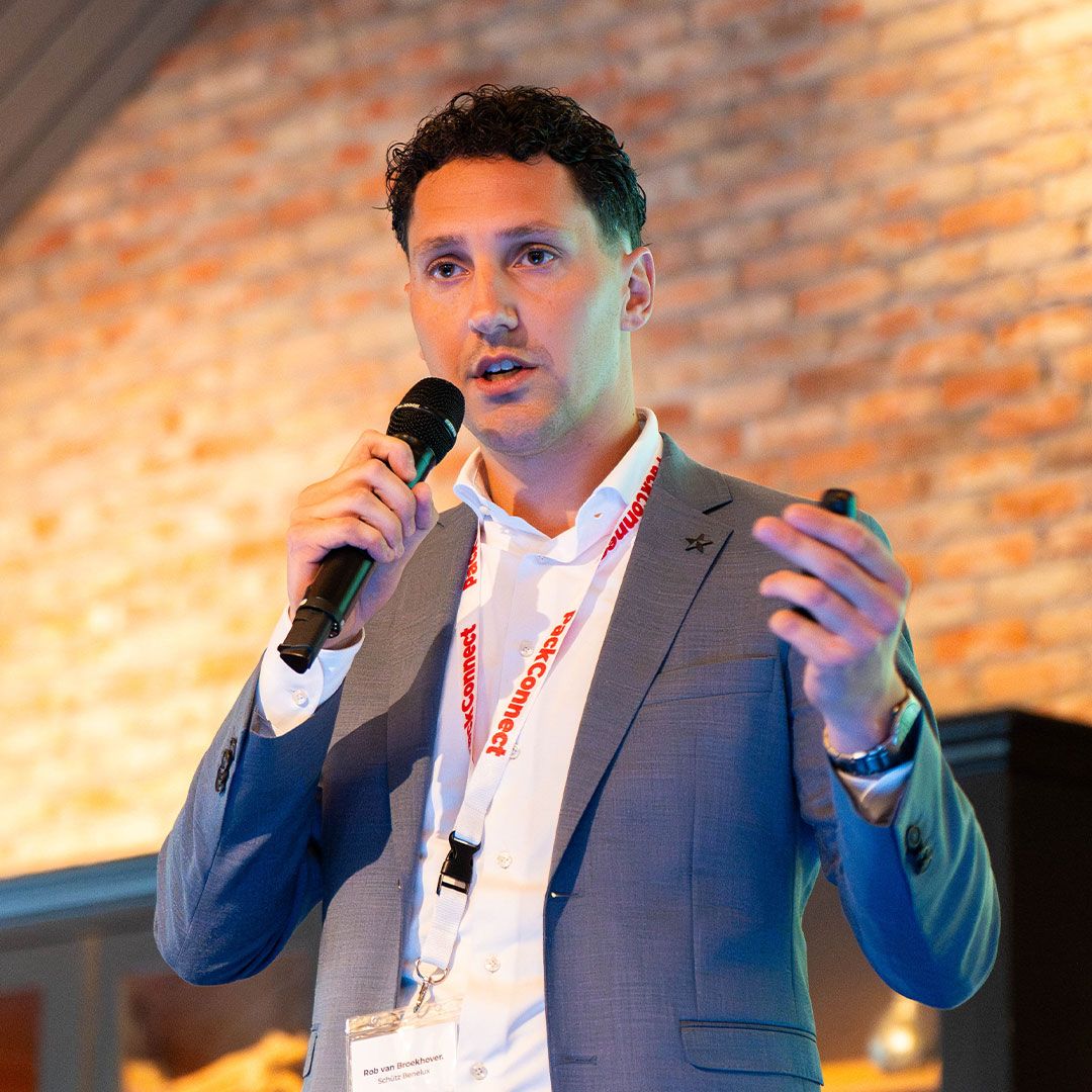 Man in a suit speaking into a microphone, wearing a lanyard, with a brick wall background.