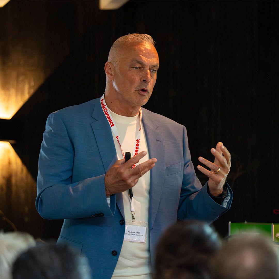 Man in a blue blazer and lanyard speaking passionately to an audience in a dimly lit room.