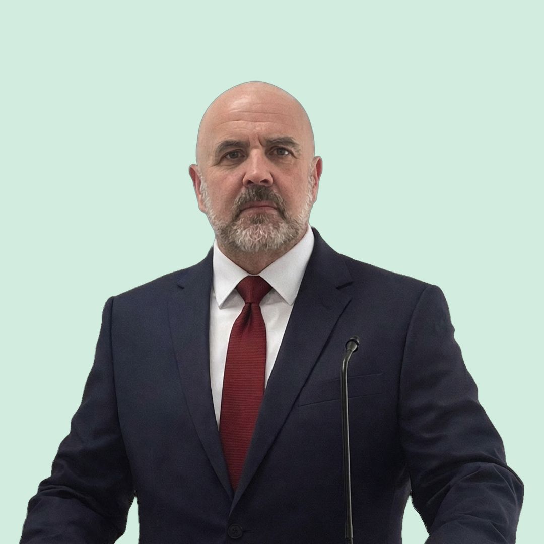 Man in a suit and red tie speaking at a podium against a light green background.