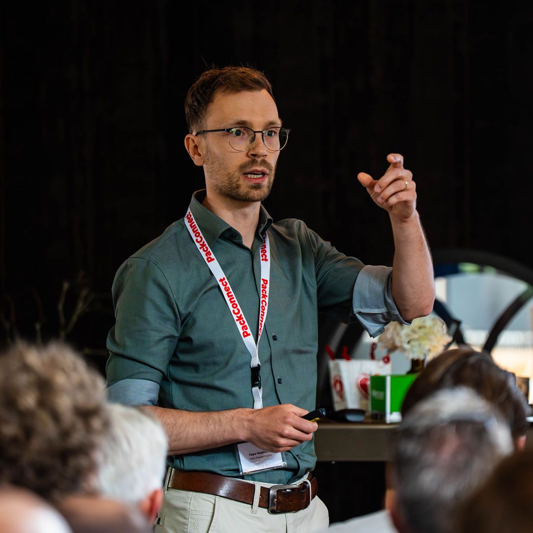 A man in glasses and a green shirt gestures while speaking at a conference, wearing a lanyard that says "PackConnect."