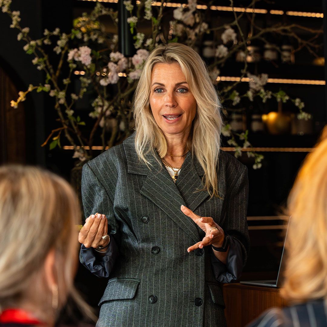 A woman with long blonde hair in a pinstripe blazer is speaking, gesturing with her hand. A laptop is beside her. Flowers are in the background.