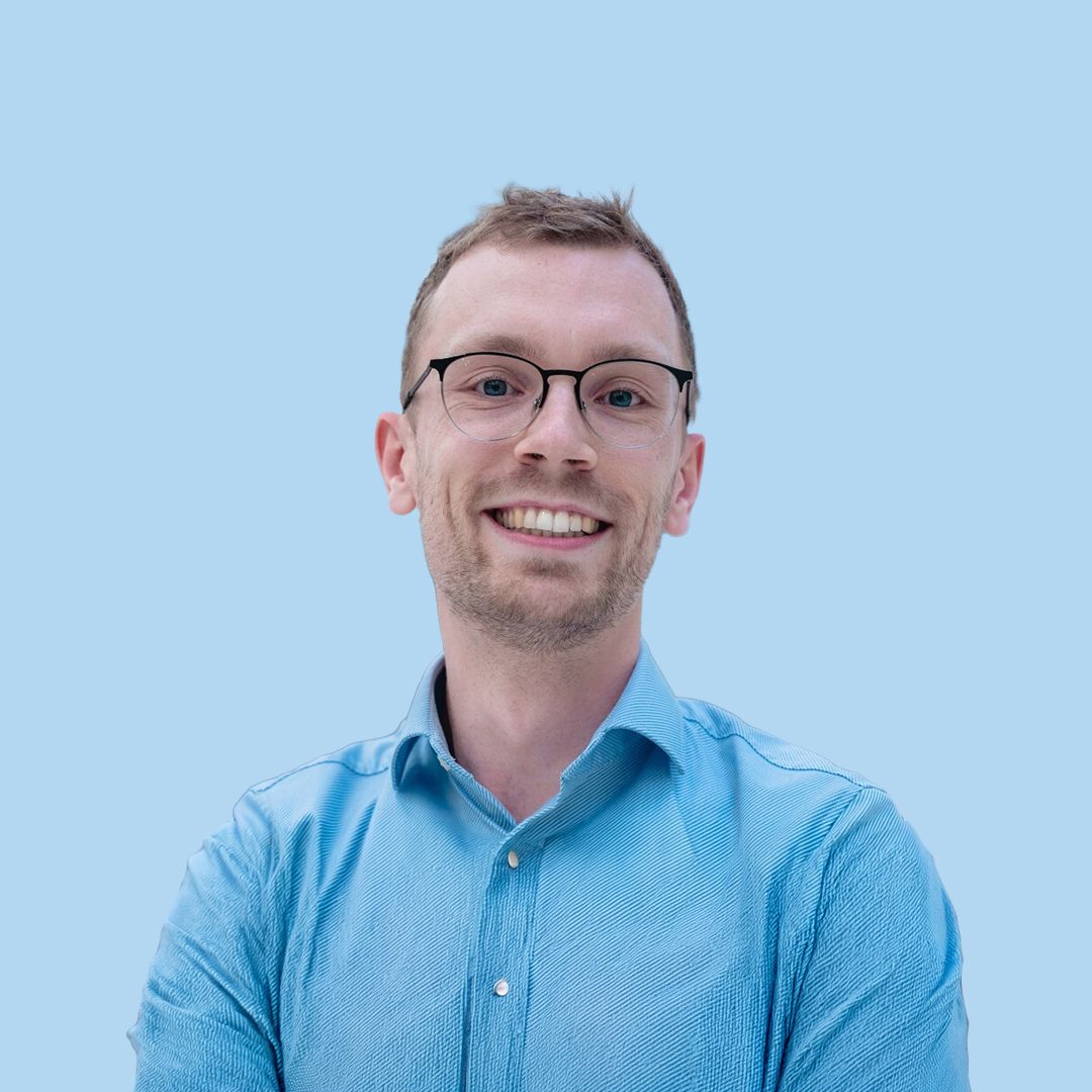 Smiling man with glasses in a light blue shirt against a pale blue background.