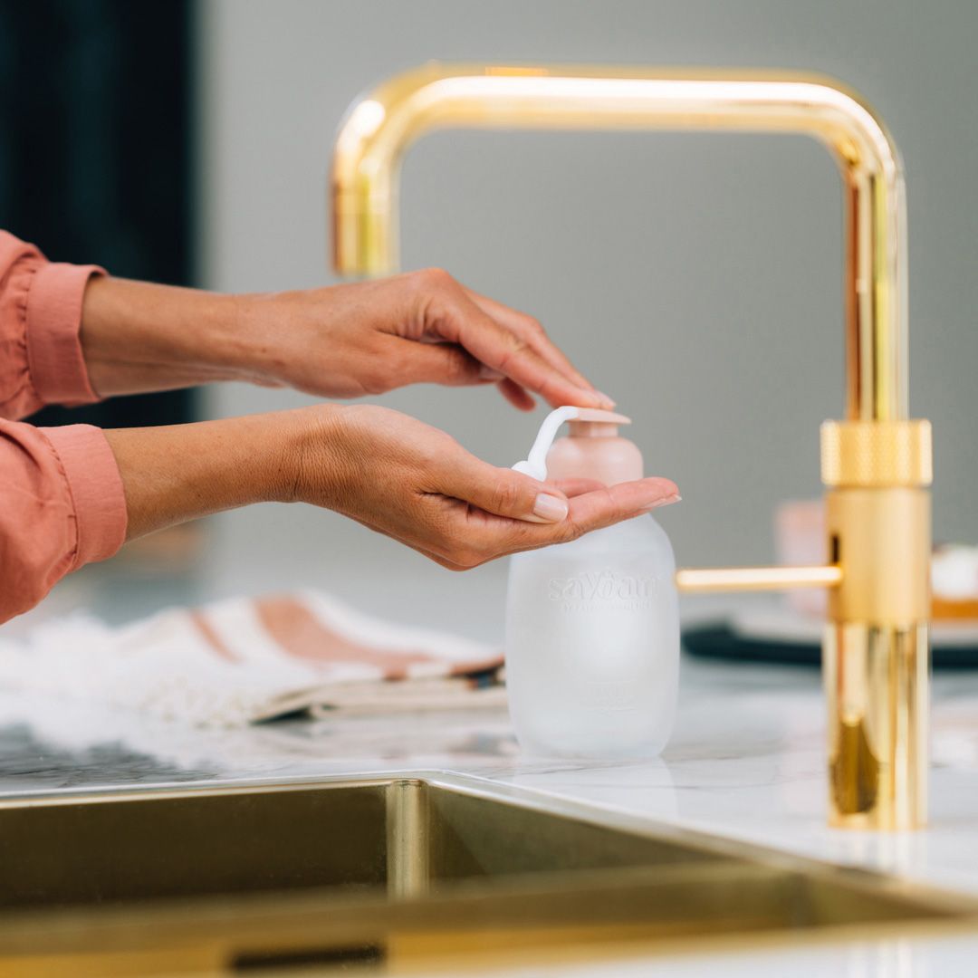 Hands pressing a soap dispenser pump beside a sleek gold kitchen faucet on a marble countertop.