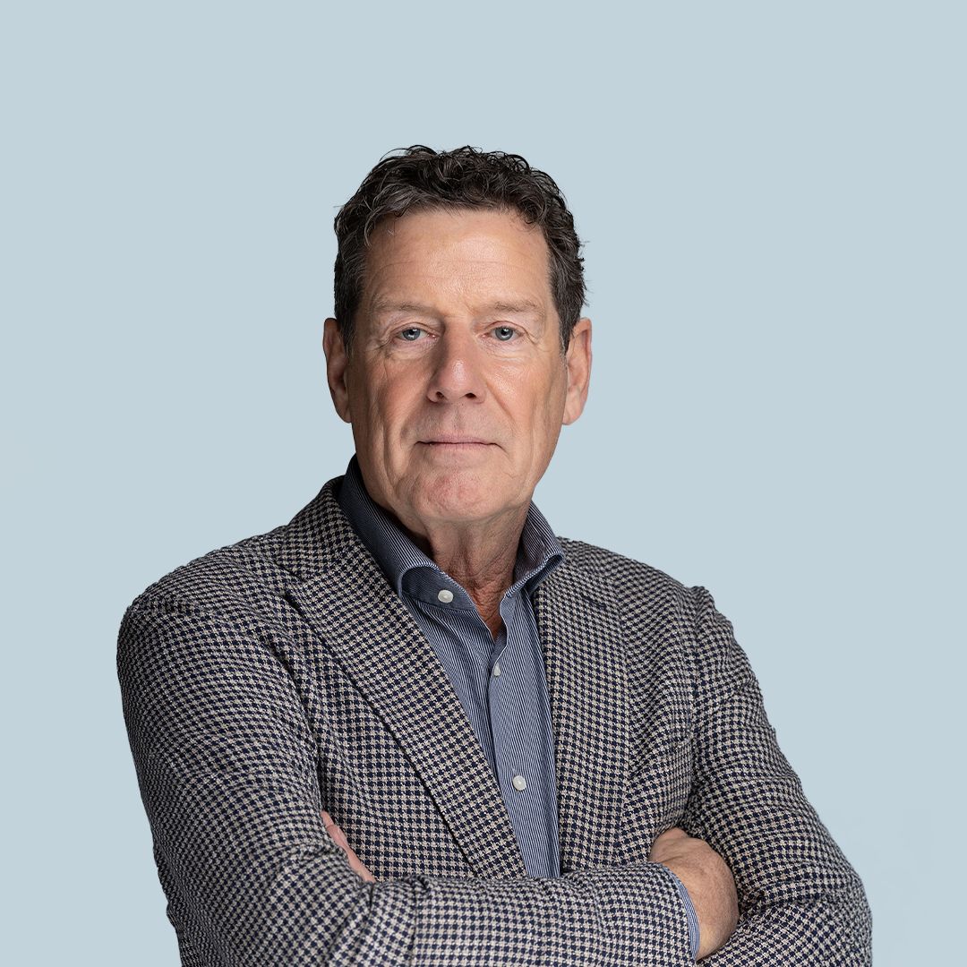 Man with short hair wearing a checked blazer and blue shirt stands with arms crossed against a light blue background.