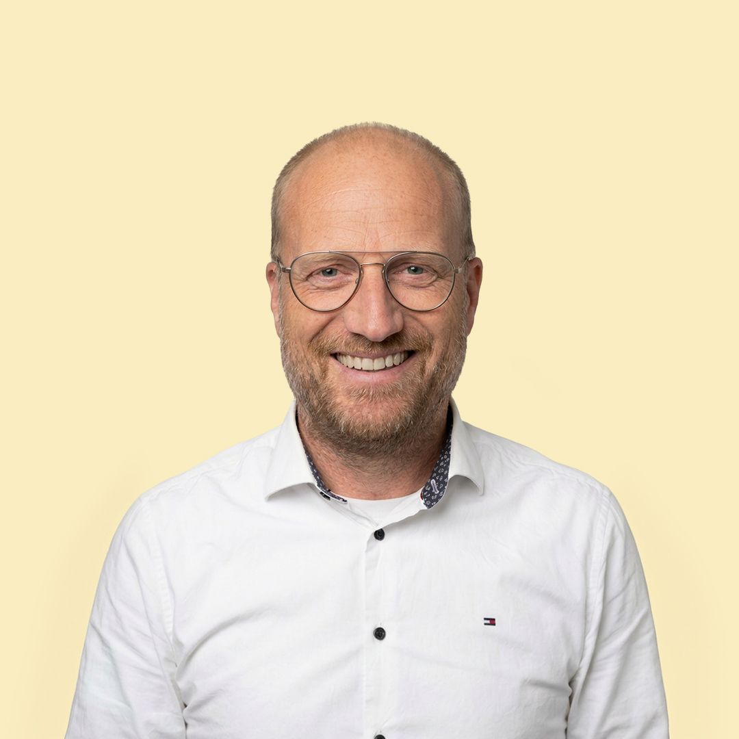 Smiling man with glasses and a beard, wearing a white shirt, against a light beige background.