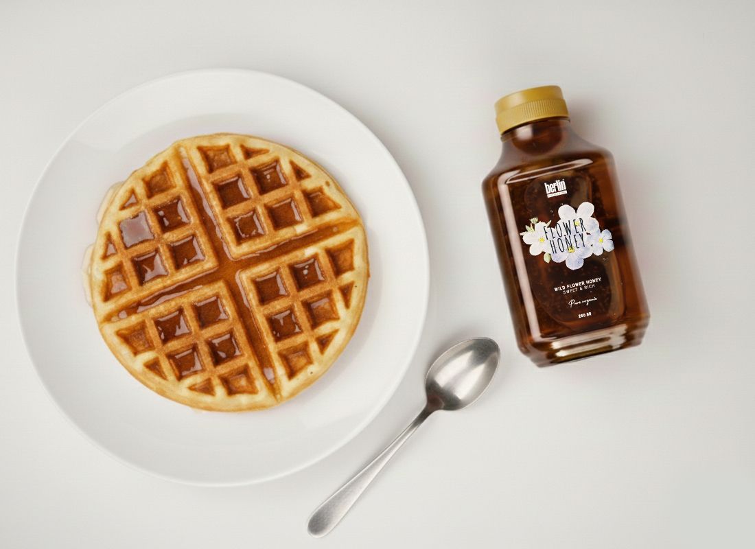 A round waffle with syrup on a white plate, next to a bottle of honey and a silver spoon on a light background.