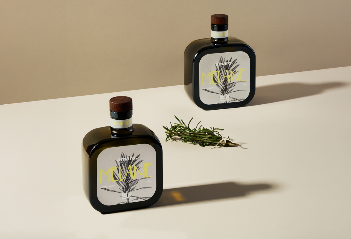 Two square-shaped brown glass bottles of amaro labeled “Melanie” with minimalist gray labels featuring botanical illustrations, placed on a light beige surface with a sprig of rosemary between them, against a neutral background.