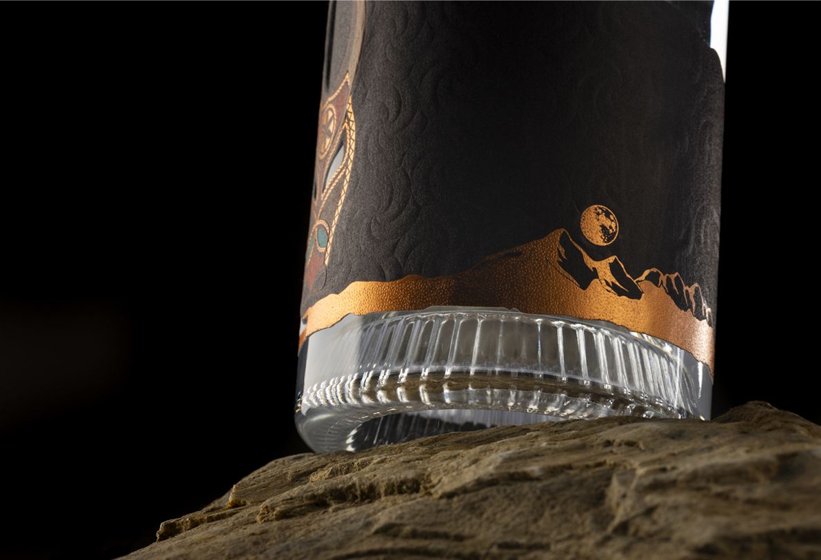 Nocturne Ribs glass bottle for spirits by Berlin Packaging customized by Redfish design agency, created for the Ginnasium project, detail of ribbed glass base and lower part of the label with debossed paper and hot stamping elements, placed on a rock surface.