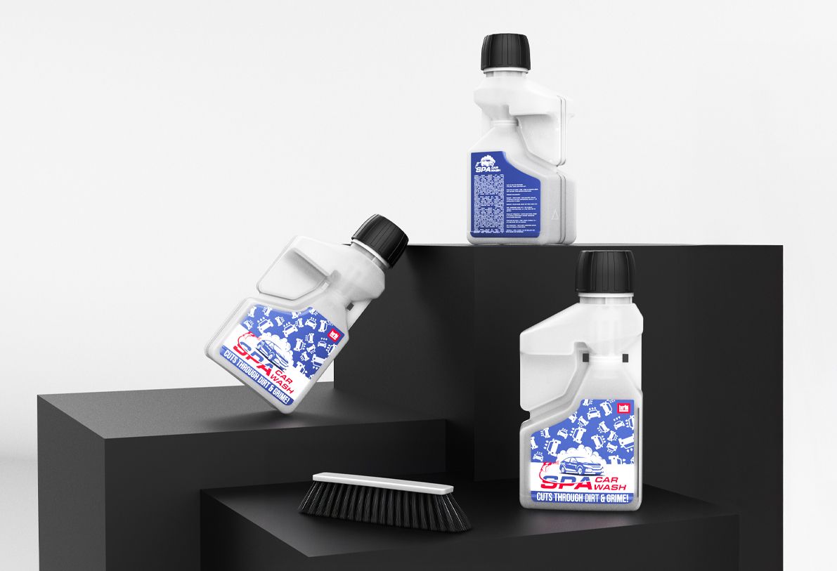 News_Dosing_bottle Three bottles of car wash solution on black steps, with a black cleaning brush nearby. Bottles feature blue and white labels.