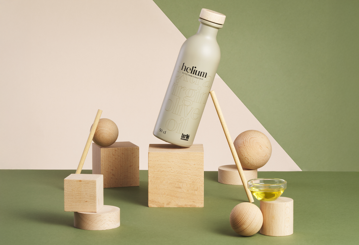 Artistic arrangement of Helium olive oil bottles in sustainable packaging with wooden geometric shapes on a green and beige background.