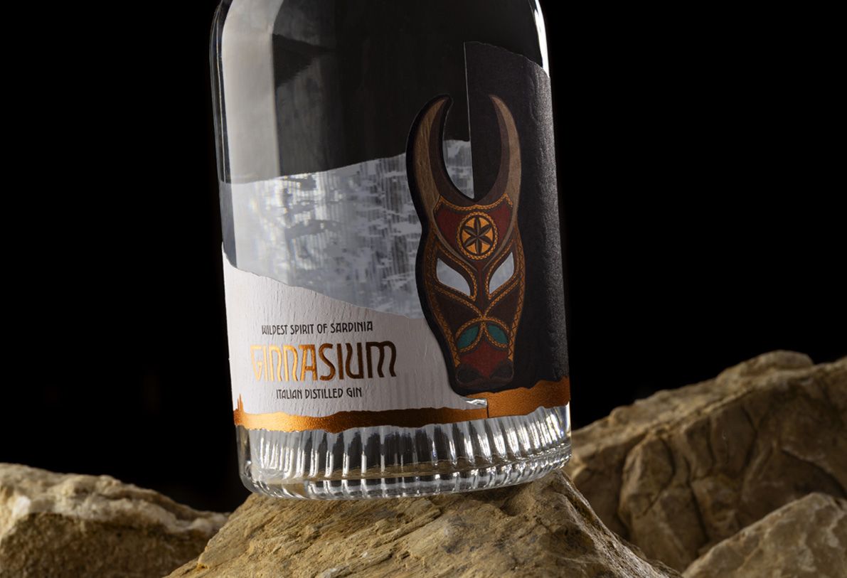 Nocturne Ribs glass bottle for spirits by Berlin Packaging customized by Redfish design agency, created for the Ginnasium project, close-up of shaped label with graphic mask illustration and hot stamping elements above ribbed glass base, positioned on rock surface.