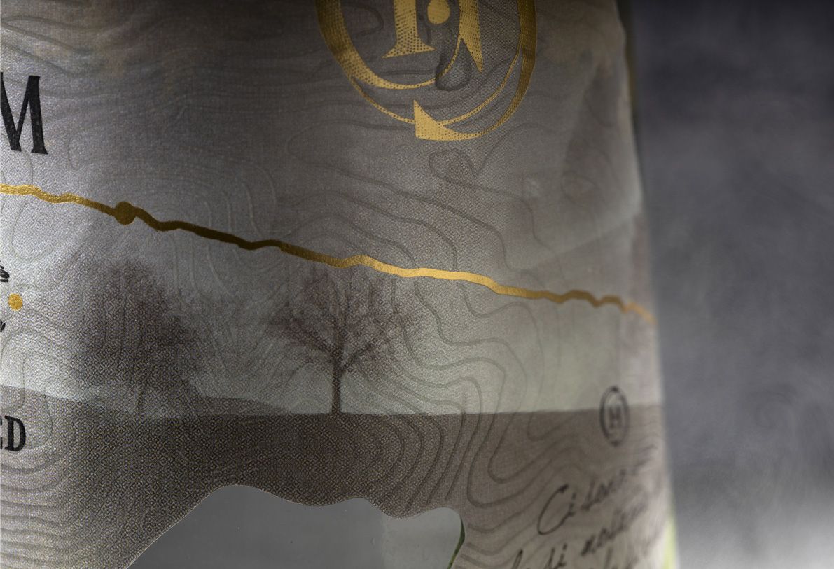 Alchemist glass bottle for spirits by Berlin Packaging customized by Rewot creative agency, created for the Ginnasium project, close-up of shaped, textured label with hot stamping elements and graphic illustration.