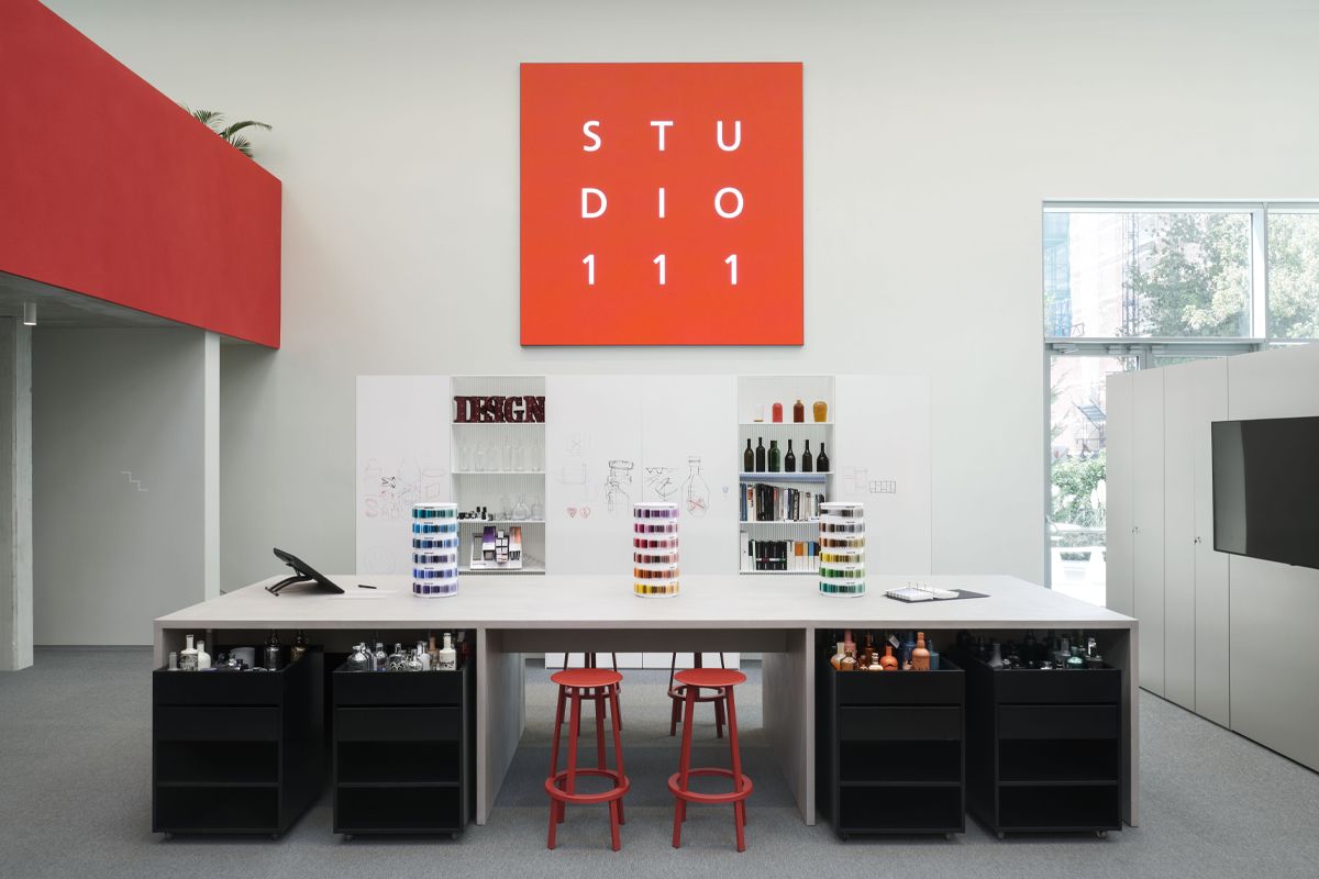 Modern studio room with a large worktable, red stools, shelves displaying various bottles, and a prominent "STUDIO 111" sign on the wall.