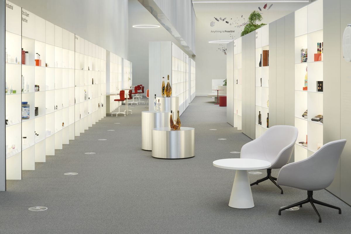 Modern office space with white shelves displaying various products, red chairs near desks, and a seating area with two gray chairs and a white table.