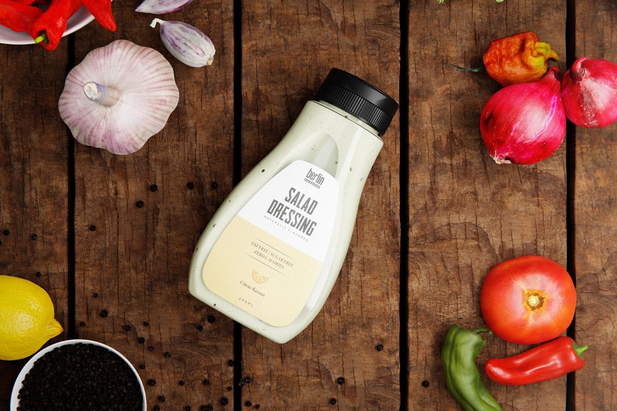 A bottle of “Salad Dressing” lies on a rustic wooden surface surrounded by fresh vegetables, including garlic, tomatoes, peppers, and a lemon, along with a bowl of black peppercorns.
