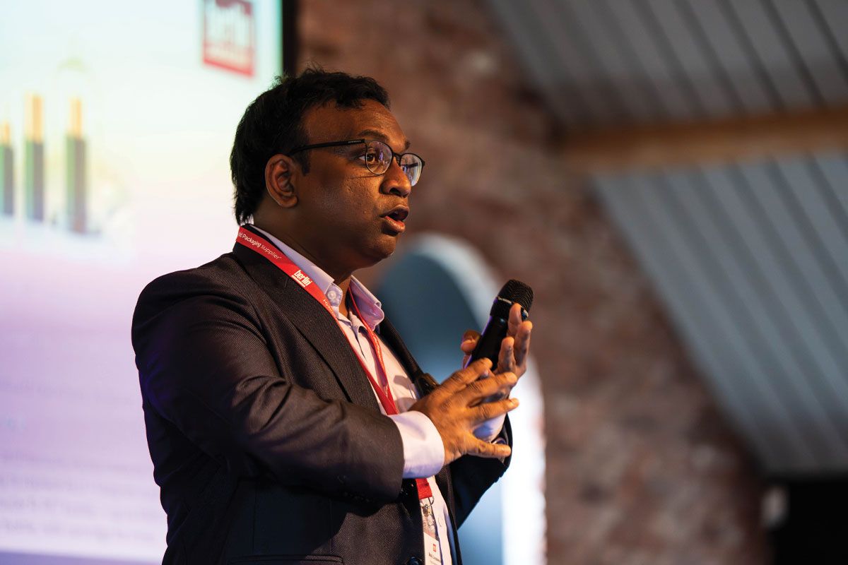 A person speaking into a microphone on stage, with a presentation slide in the background.