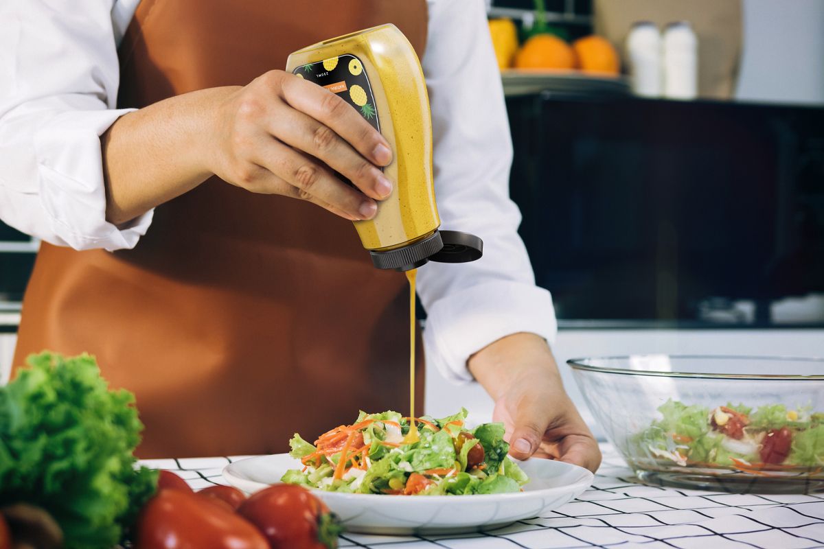 A person wearing a brown apron is pouring yellow dressing or sauce from a squeeze bottle onto a fresh salad in a white bowl. Fresh vegetables and another bowl of salad are on the counter nearby.