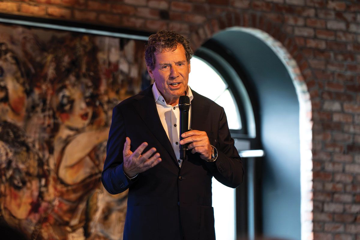 A person in a suit speaks into a microphone in front of a brick wall with a vibrant abstract painting in the background.