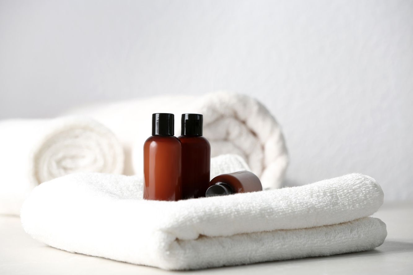 Two brown bottles on stacked white towels, with a rolled towel in the background, set against a plain white backdrop.