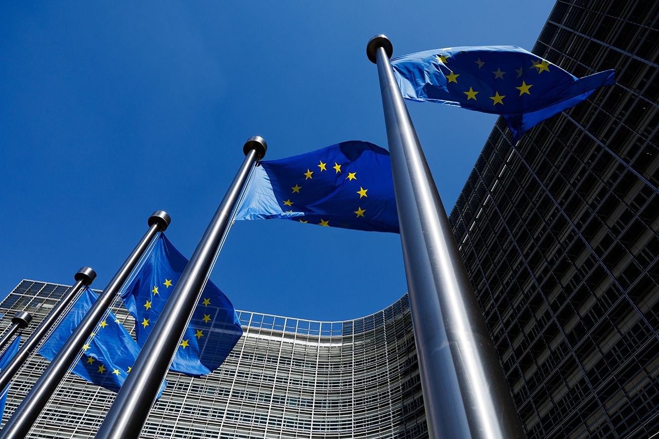 European Union flags waving on tall poles in front of a modern glass building.