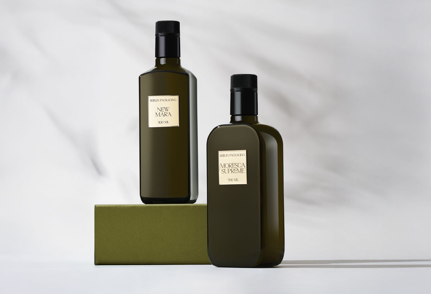 Originae Collection olive oil bottles by Berlin Packaging, featuring two premium dark green glass bottles with black caps and metallic gold labels. Labels read "New Mara 250 ml" and "Moresca Supreme"'. Minimalist composition on a light background with a green block