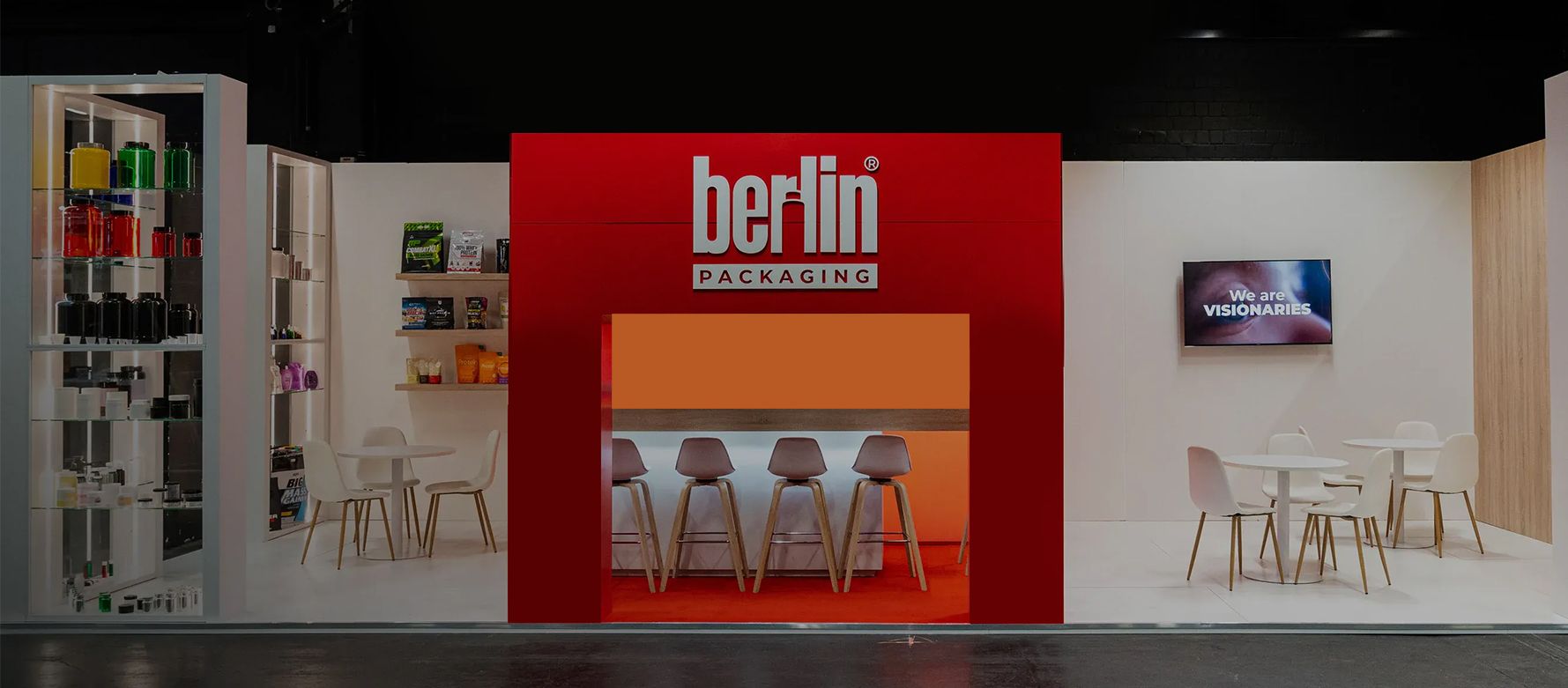 Exhibition booth with "berlin packaging" sign, red backdrop, white chairs, tables, and a display shelf with colorful bottles.