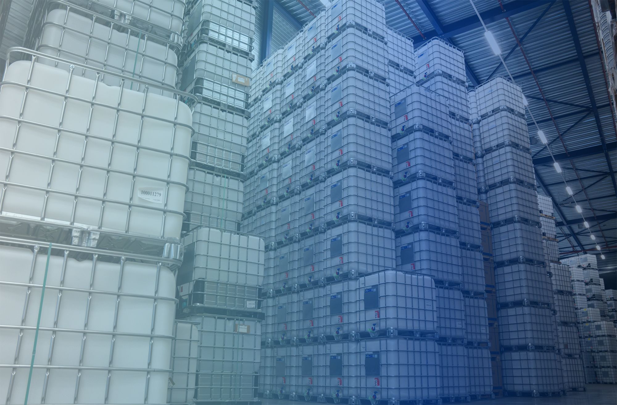 Stacks of large, cubical industrial containers in a spacious warehouse with metal roofing and fluorescent lighting.