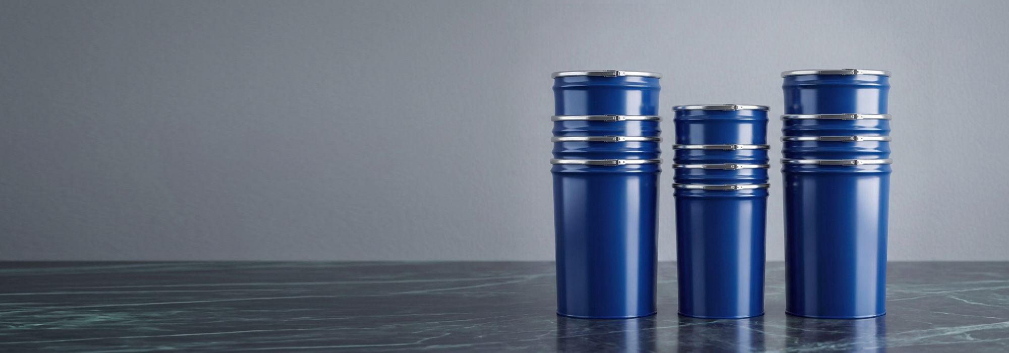 Three blue metal containers with silver bands stand on a dark marble surface against a gray background.