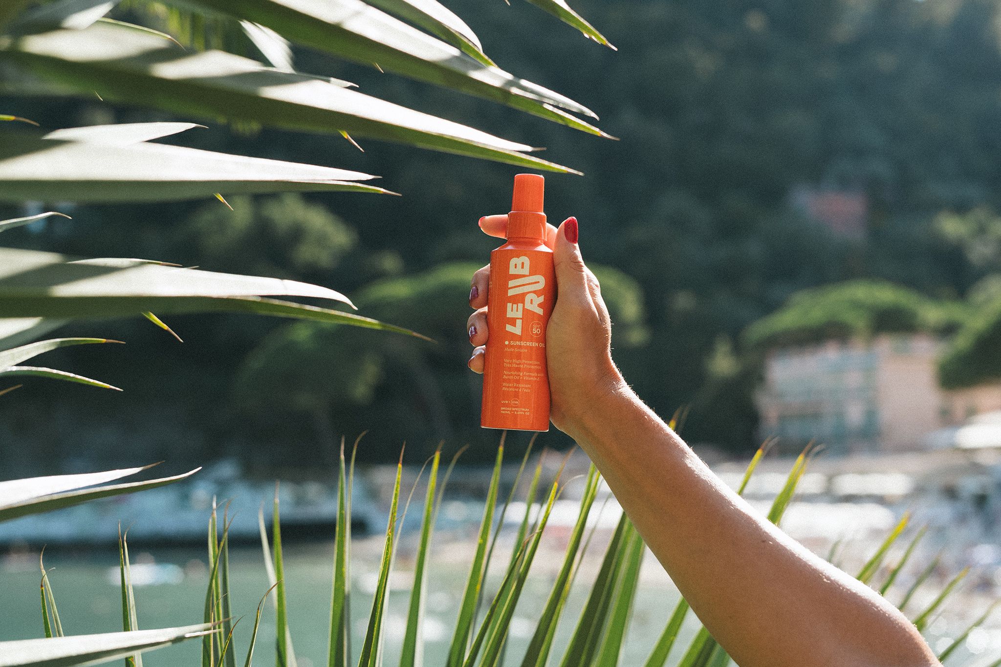 Le Rub sunscreen Oil A hand holding an orange bottle of “LE RUB” sunscreen oil among green palm leaves, with a coastal landscape and water visible in the background.