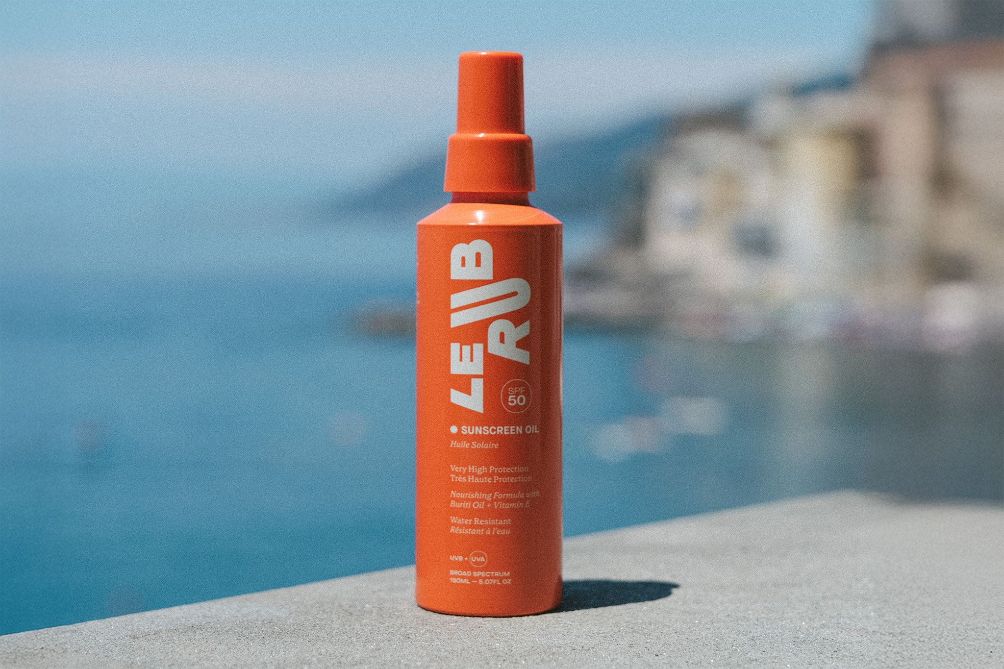Le rub sunscreen oil A hand holding a bright orange bottle of “LE RUB” sunscreen oil against a backdrop of clear blue sky and a glimpse of the ocean.