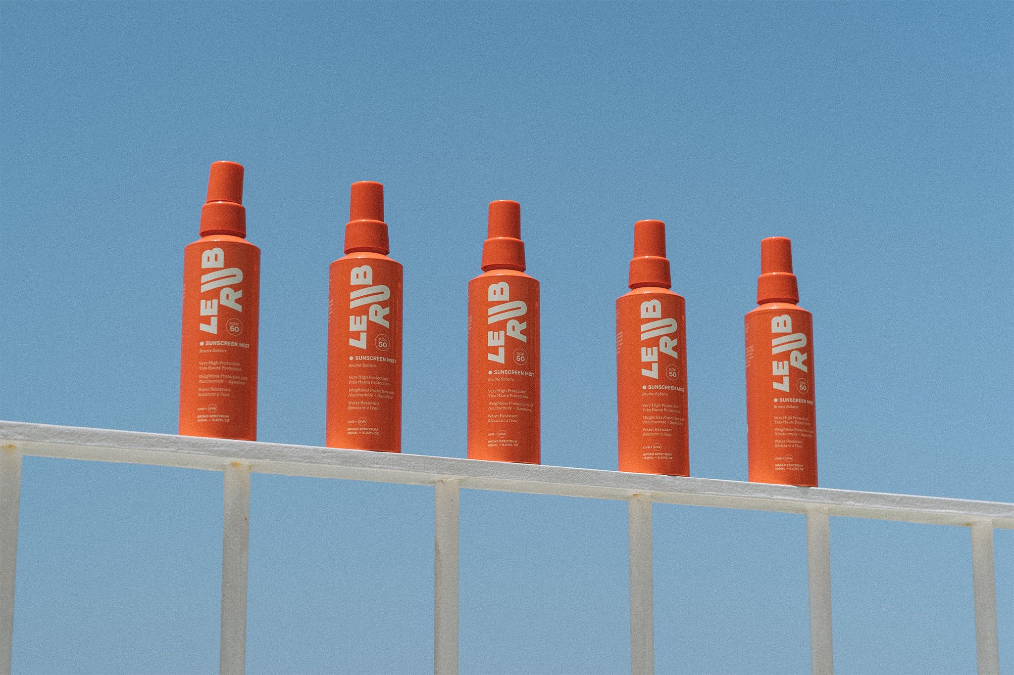 Four bright orange bottles of “LE RUB” sunscreen oil lined up on a white railing, with a deep blue ocean and clear sky in the background.