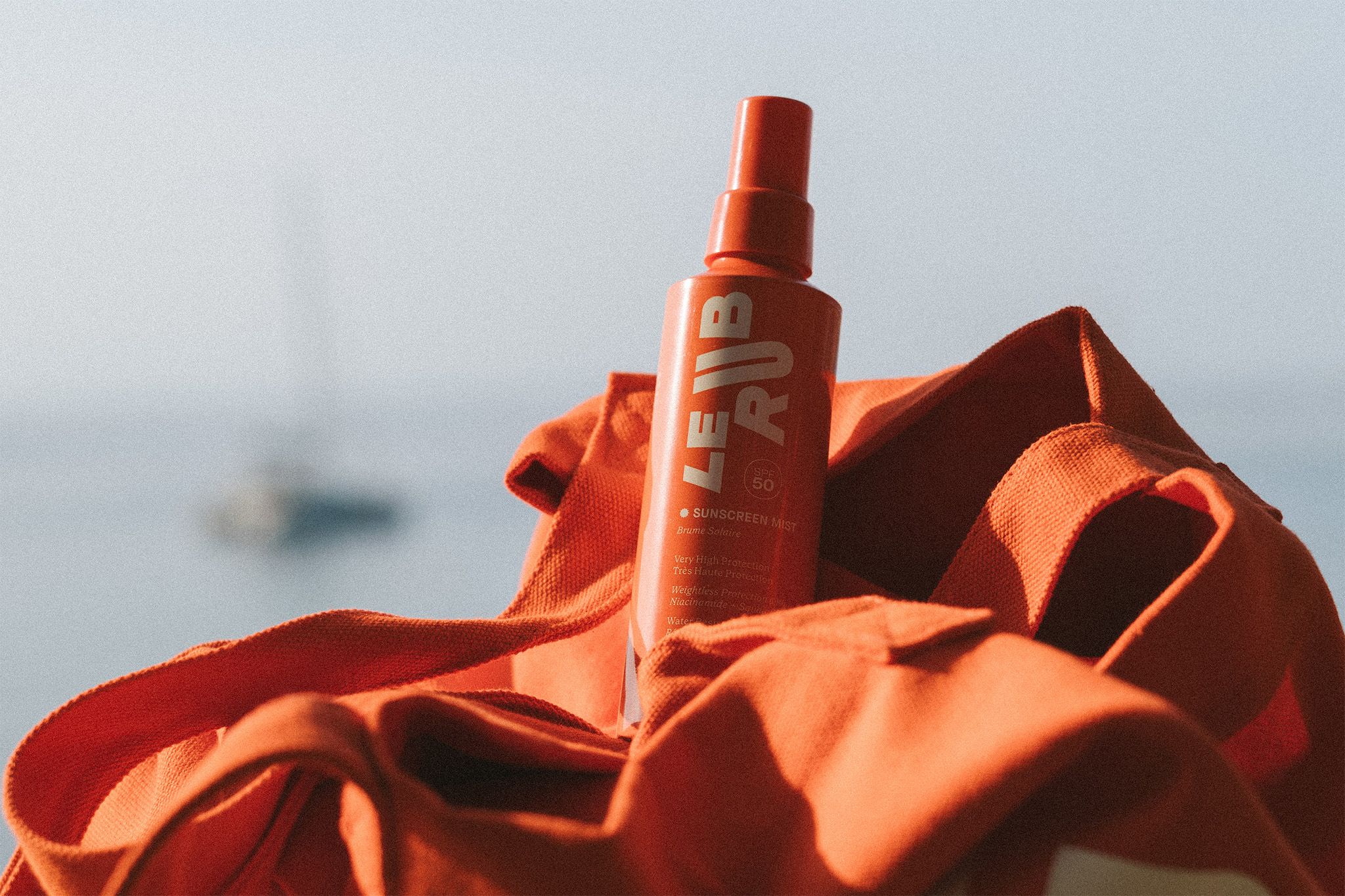 Le Rub sunscreen Mist An orange bottle of “LE RUB” sunscreen mist resting on an orange beach bag, with a blurred sailboat and calm sea in the background.
