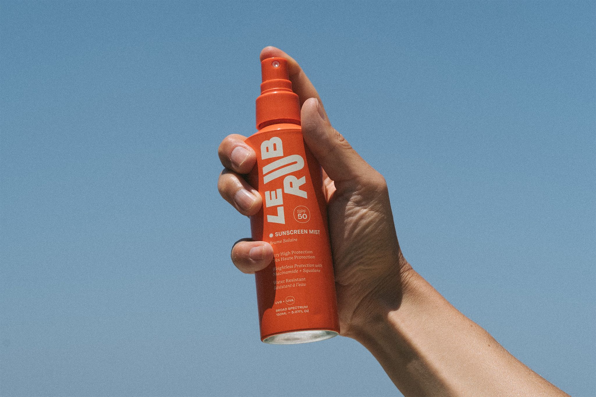 Le Rub sunscreen Mist A hand holding a bright orange bottle of “LE RUB” sunscreen oil against a backdrop of clear blue sky and a glimpse of the ocean.