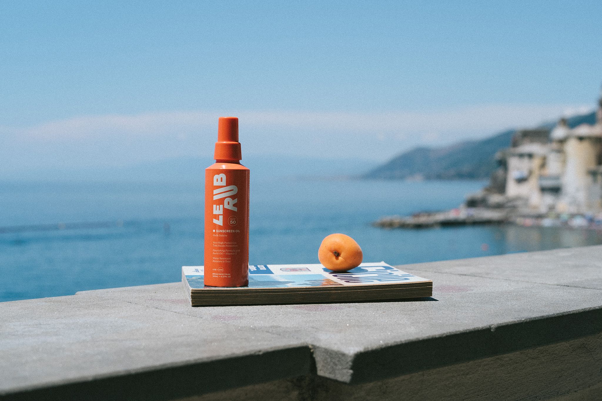 Le Rub sunscreen Oil An orange bottle of “LE RUB” sunscreen oil placed on a magazine next to an apricot, with a scenic view of the sea and coastal buildings in the background under a clear blue sky.