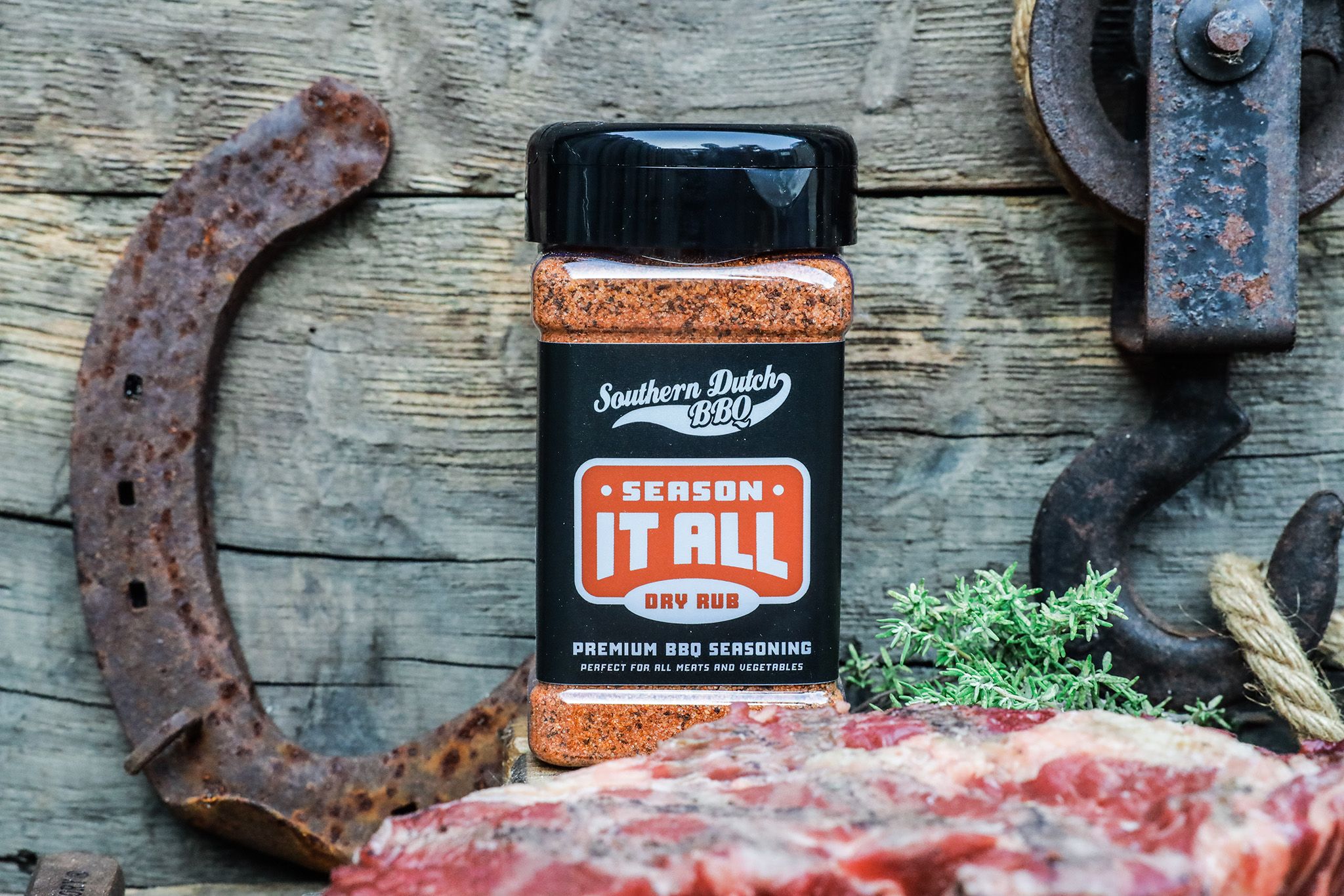 A bottle of "Season It All" BBQ dry rub against a rustic wooden background, with rusty horseshoes, herbs, and raw meat in the foreground.