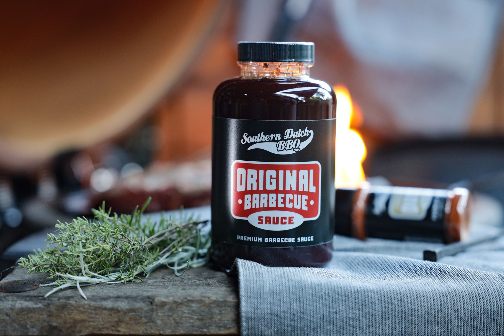 A jar of Southern Dutch BBQ Original Barbecue Sauce on a cloth-covered table with fresh herbs and a blurred background featuring a flame.