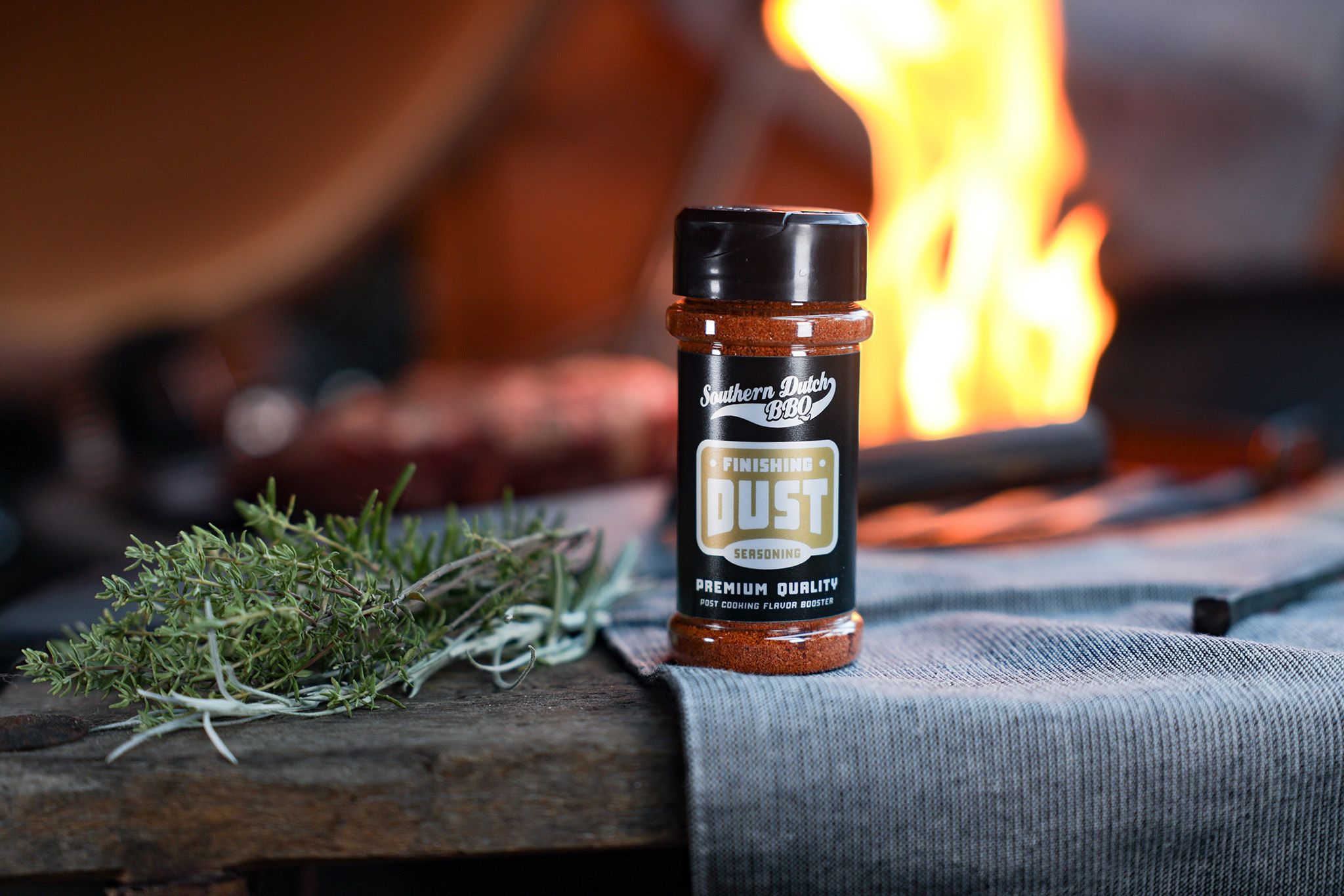 A bottle of "Finishing Dust" seasoning on a wooden surface with fresh herbs and a warm fire in the background.