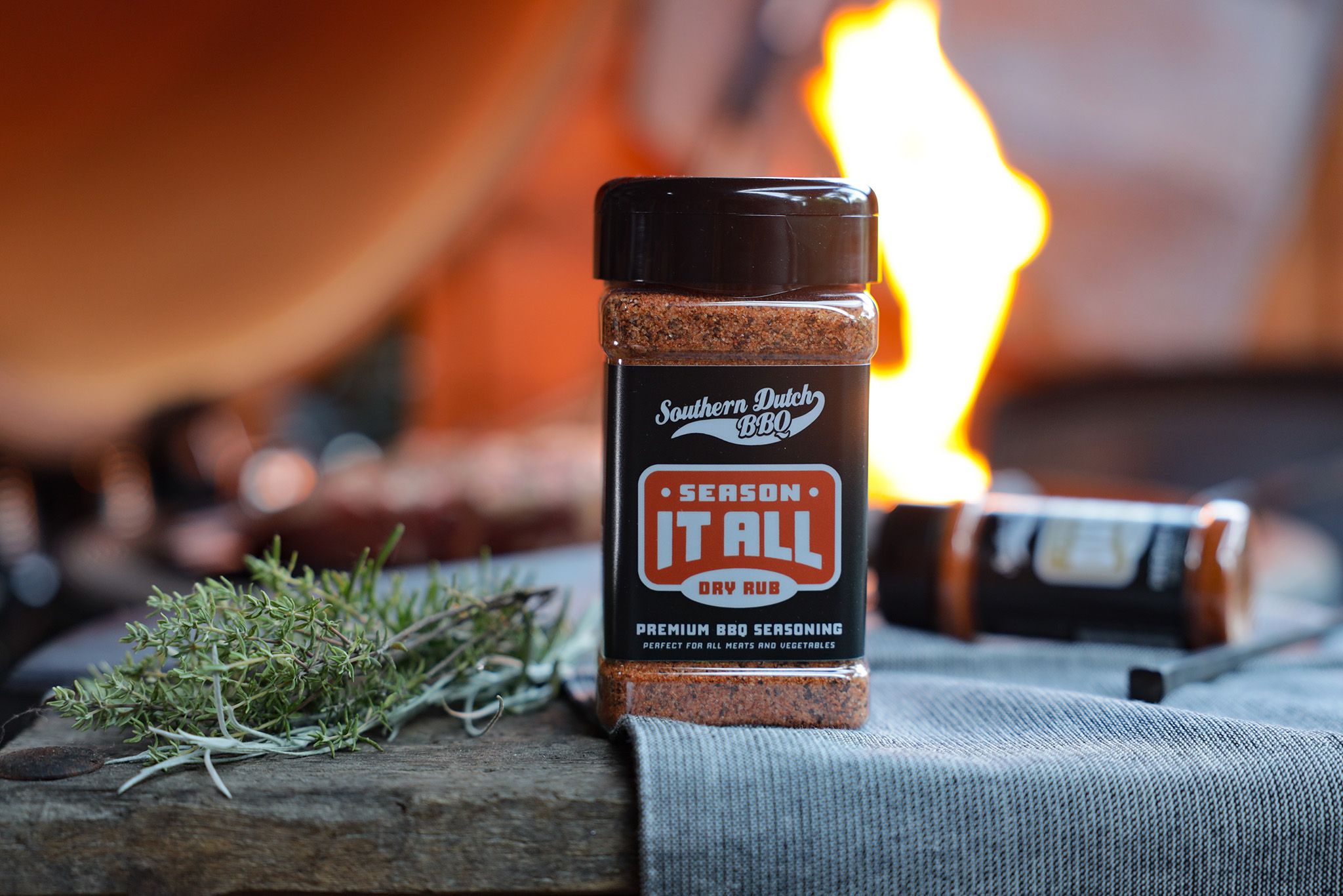 A jar of Southern Dutch BBQ "Season It All" dry rub sits on a wooden surface with fresh herbs, while a grill fire blazes in the background.