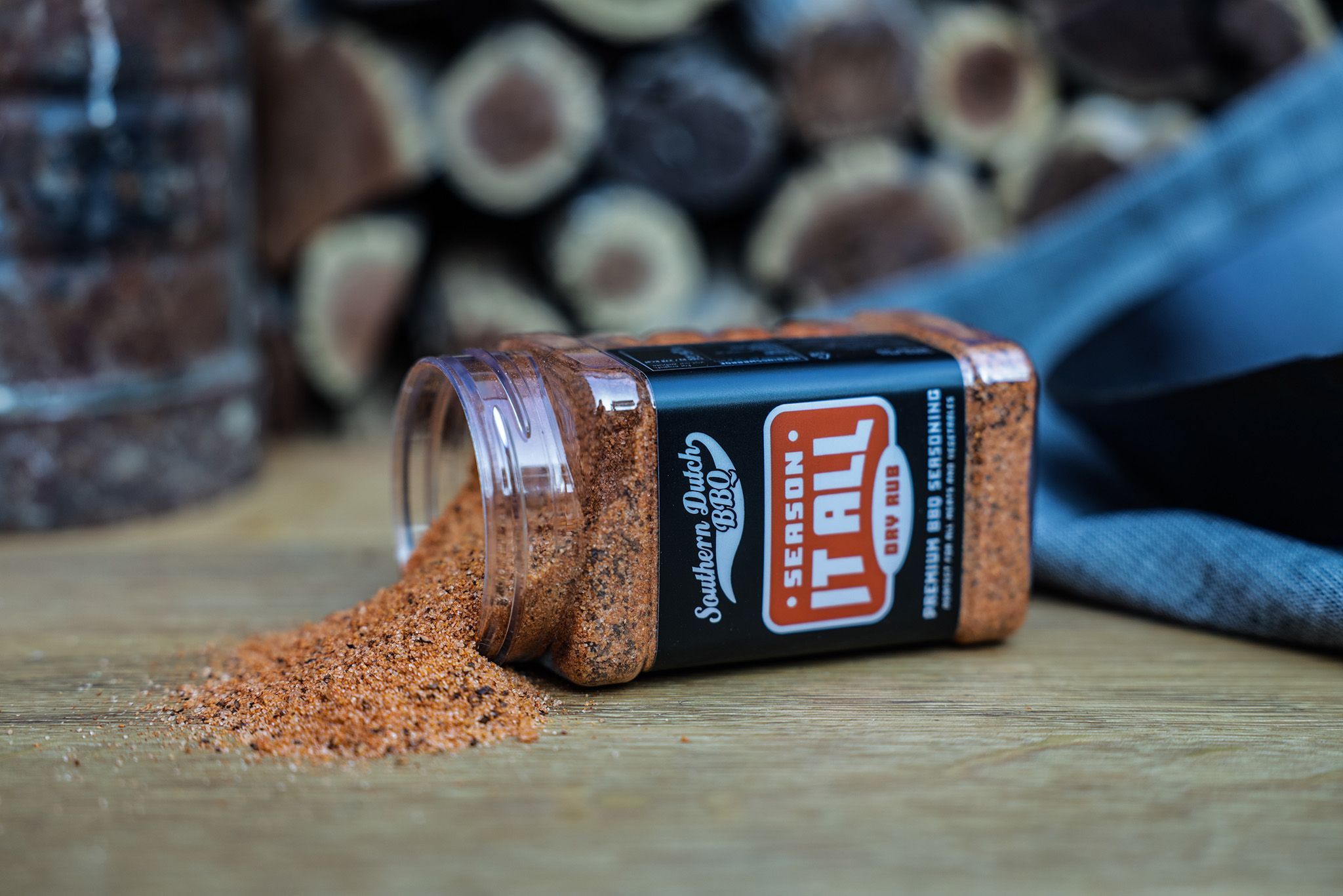 A jar of "Season It All" dry rub seasoning is tipped over, spilling orange spices onto a wooden surface. Logs and fabric are in the background.