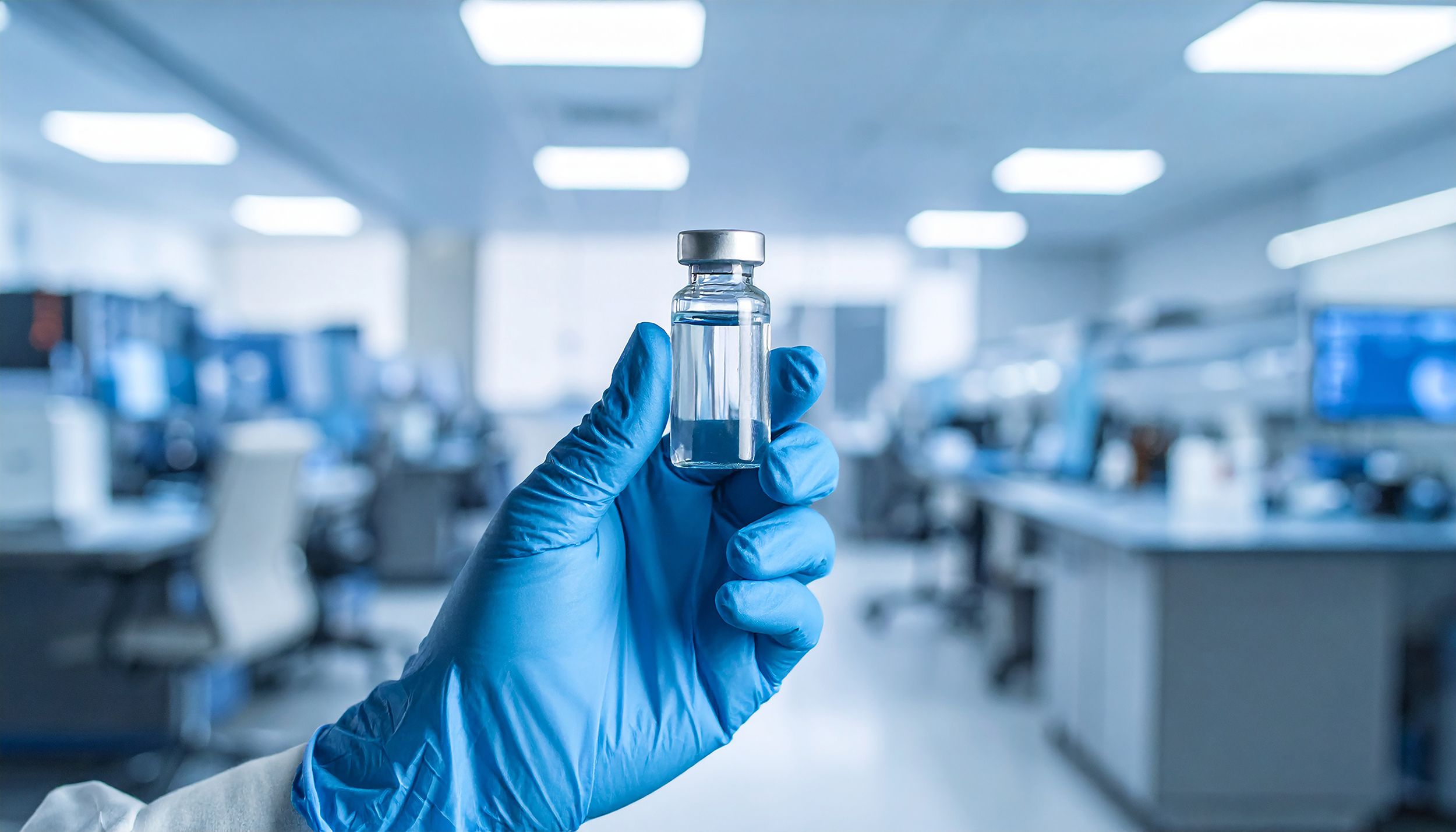 A gloved hand holding a small glass vial filled with clear liquid in a modern laboratory setting, with blurred lab equipment and workstations in the background.