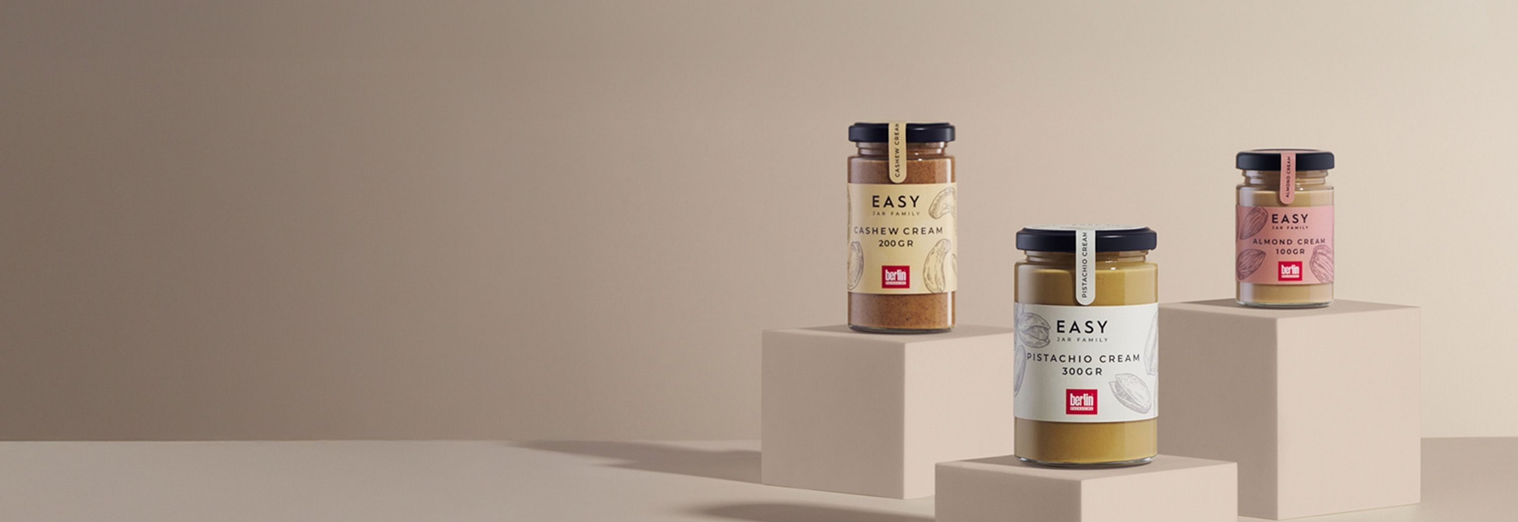 Three jars from the Easy Jar Family collection displayed on beige blocks: cashew cream (200 grams), pistachio cream (300 grams), and almond cream (100 grams).