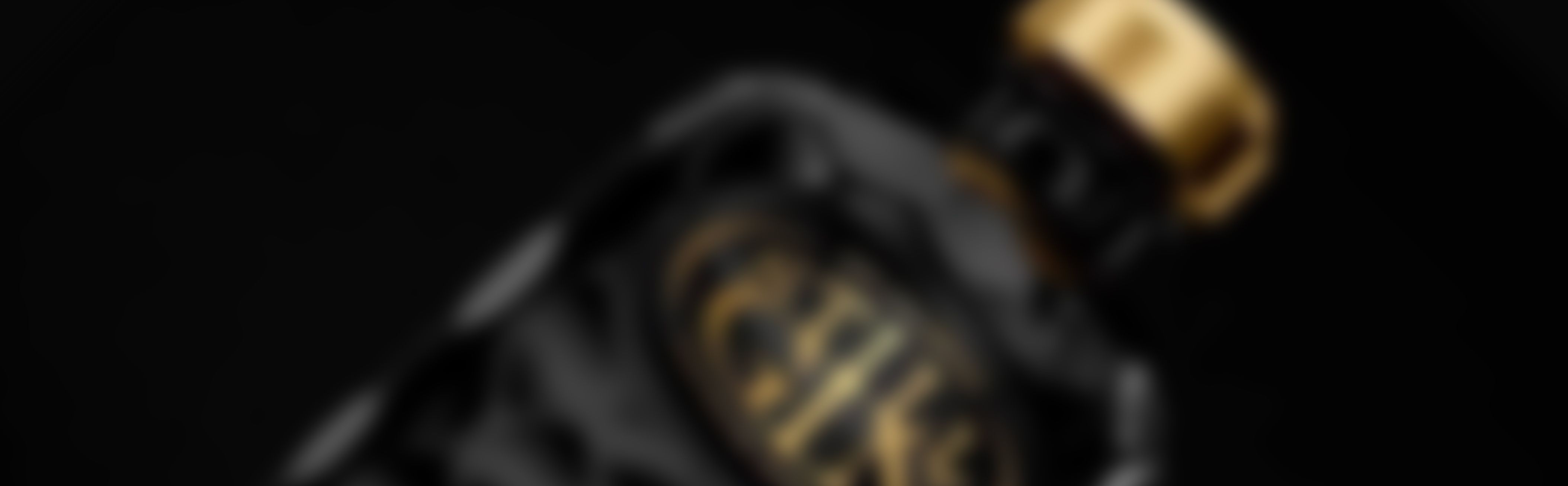 A blurred image of a black bottle with gold accents and a gold cap on a dark background.