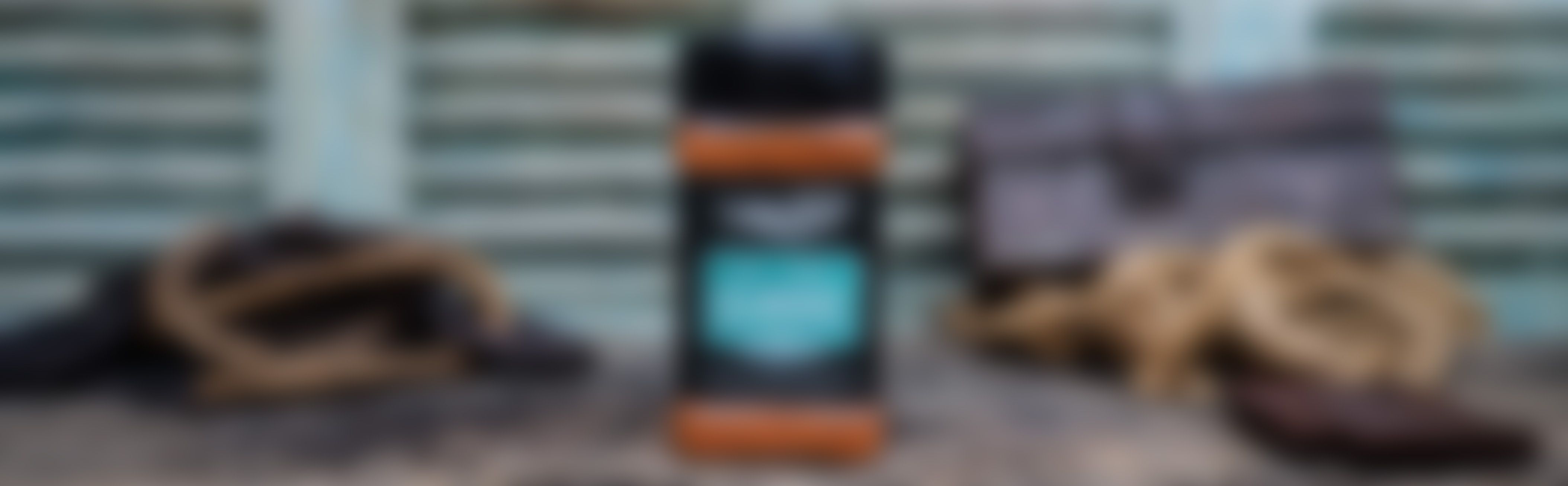 A bottle of men's body wash with orange and black packaging on a rustic wooden surface, background shows blurred accessories.