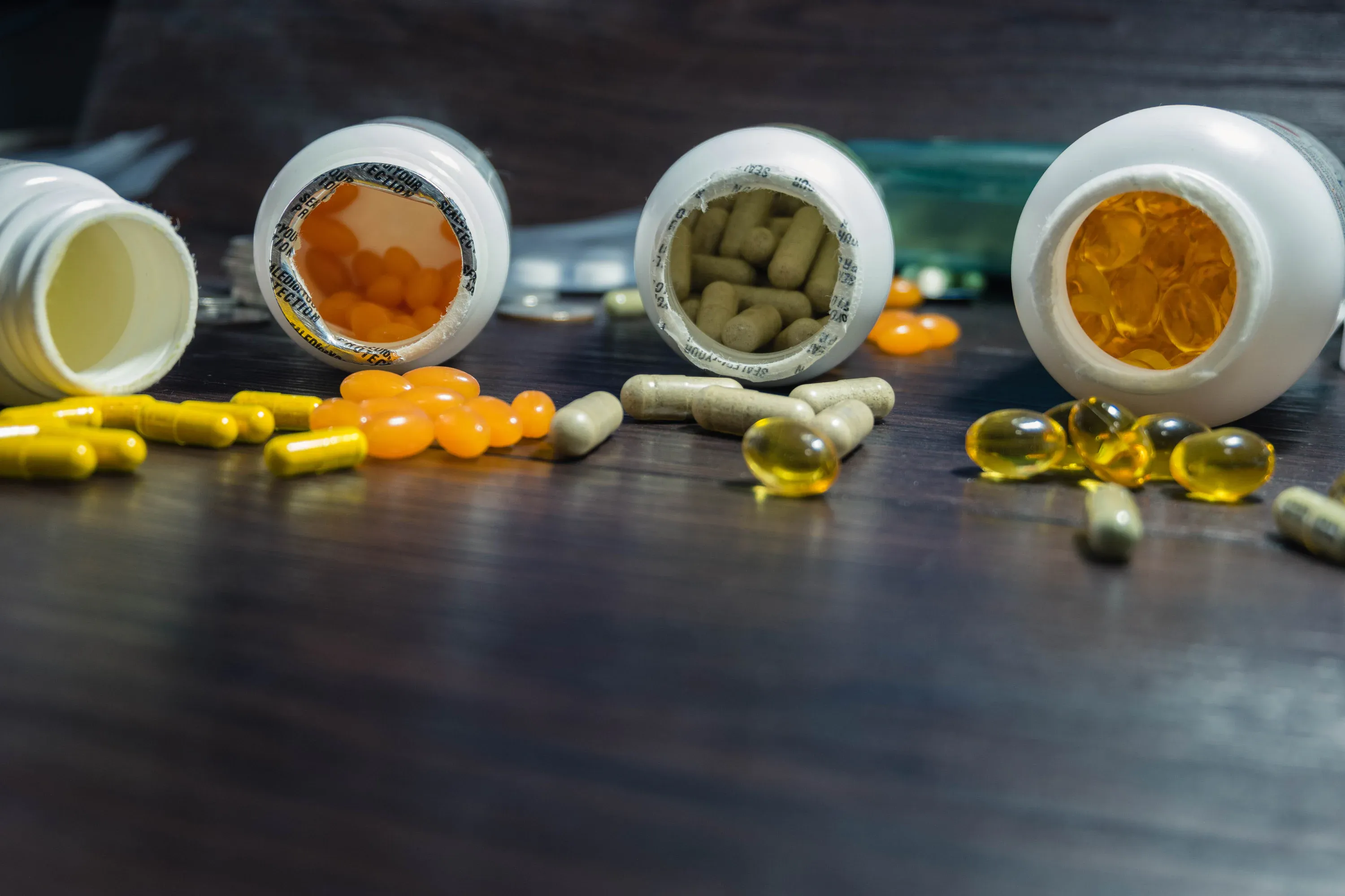 Four white supplement bottles lying on their sides with assorted capsules and softgel pills—orange, green, and yellow—scattered on a dark wooden surface for a health-focused composition.