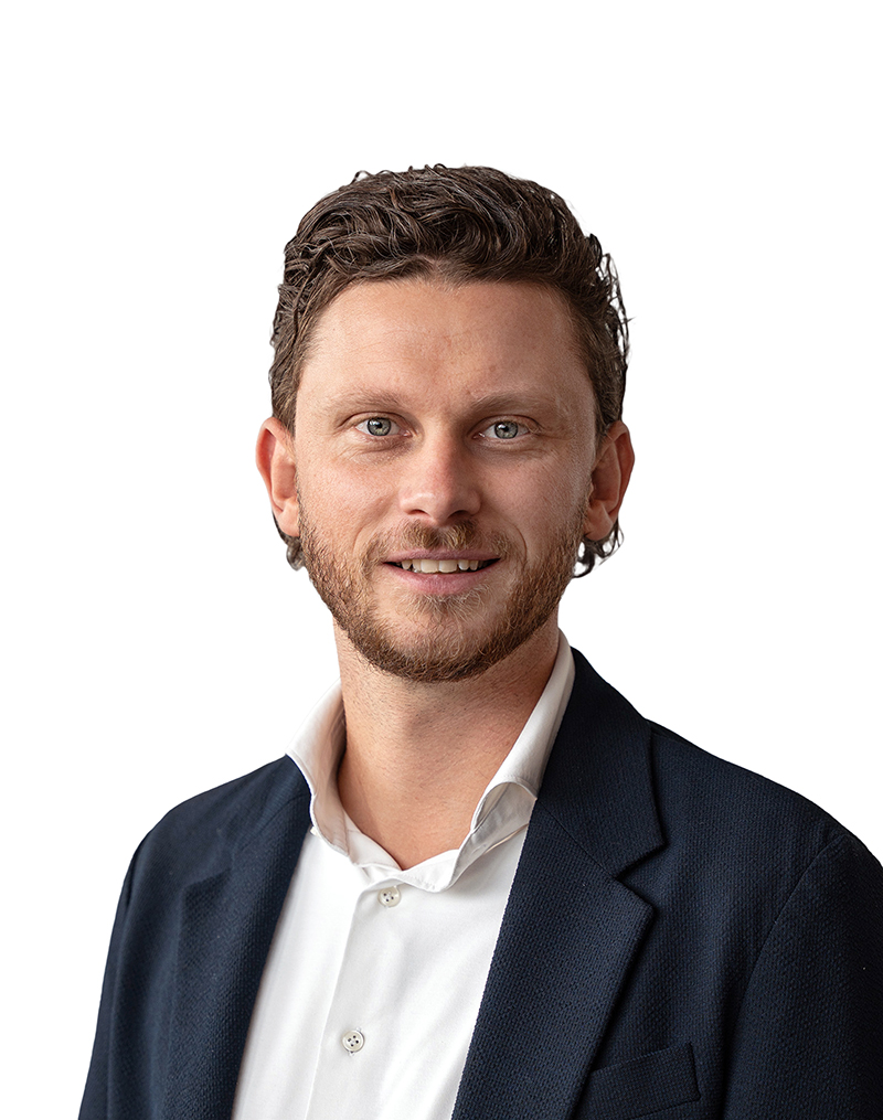 A man with short, wavy brown hair and a beard, wearing a white shirt and dark blazer, smiles against a white background.