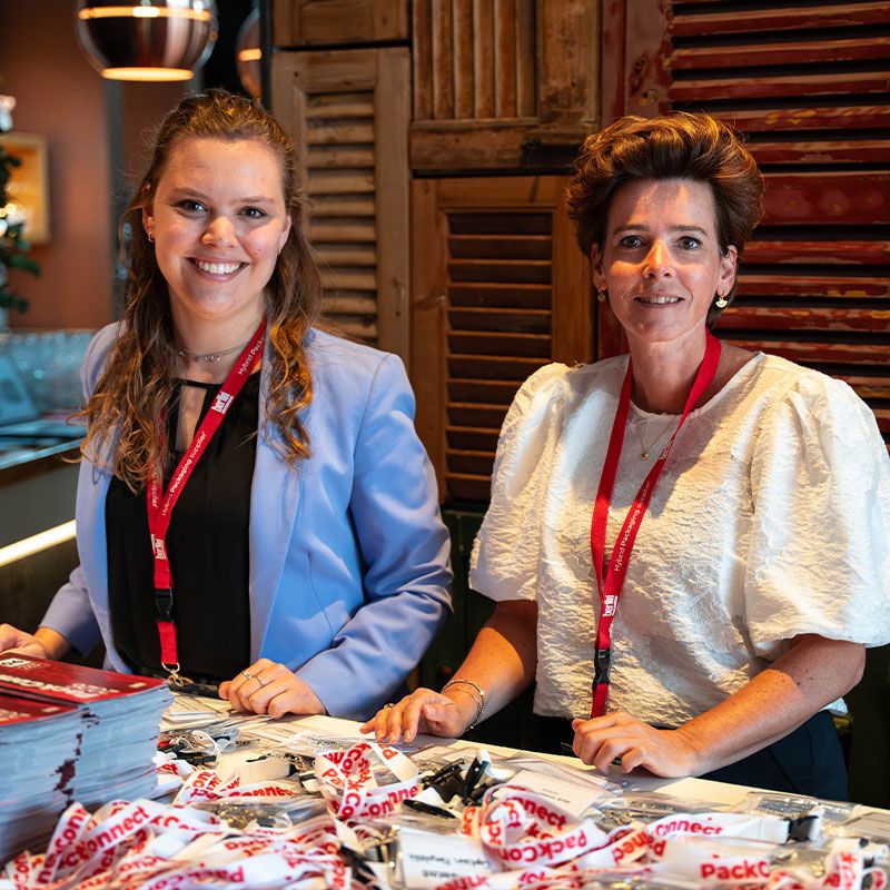 Two women wearing red lanyards stand behind a table with event materials, smiling at the camera.