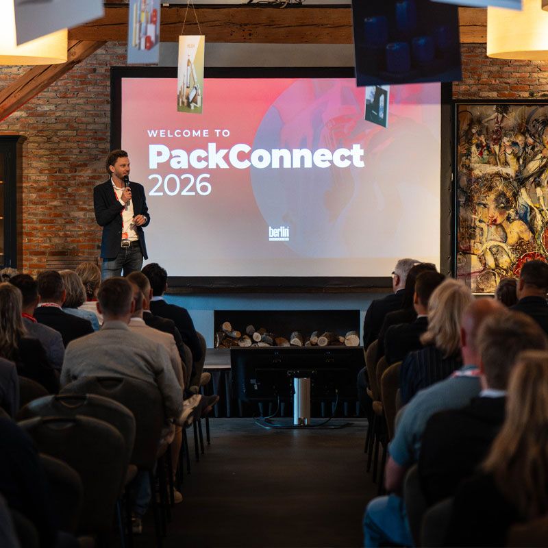 A speaker presents at the PackConnect 2026 event in Berlin, addressing a seated audience in a rustic venue with a large screen display.