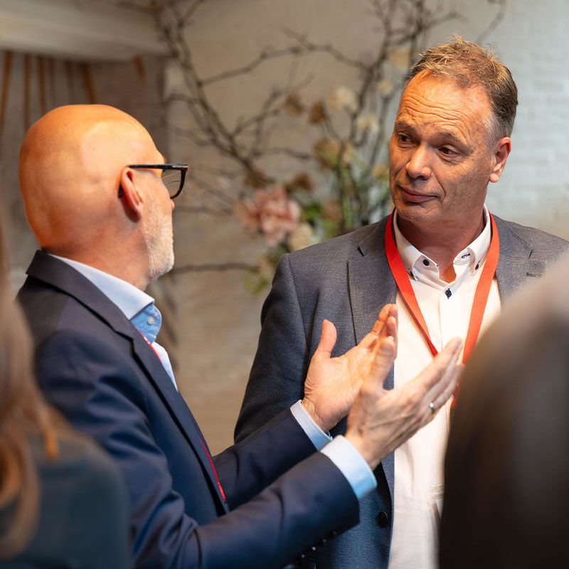 Two men in suits are having an animated discussion indoors, one gesturing with his hands. Both wear red lanyards.