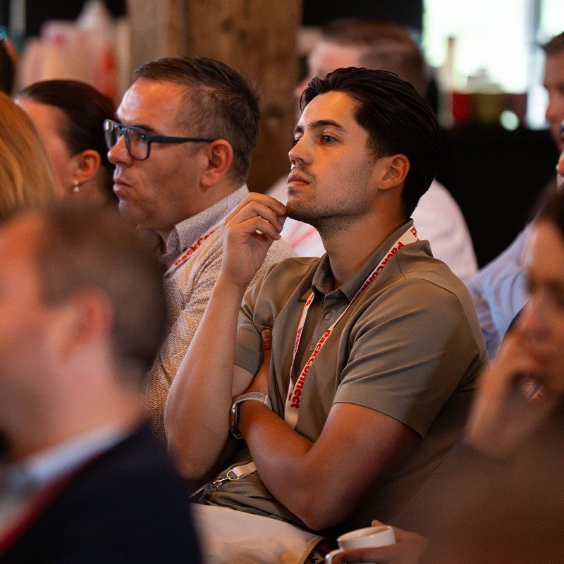 People attentively listening at a conference, wearing lanyards and sitting closely together, with a warm, focused ambiance.